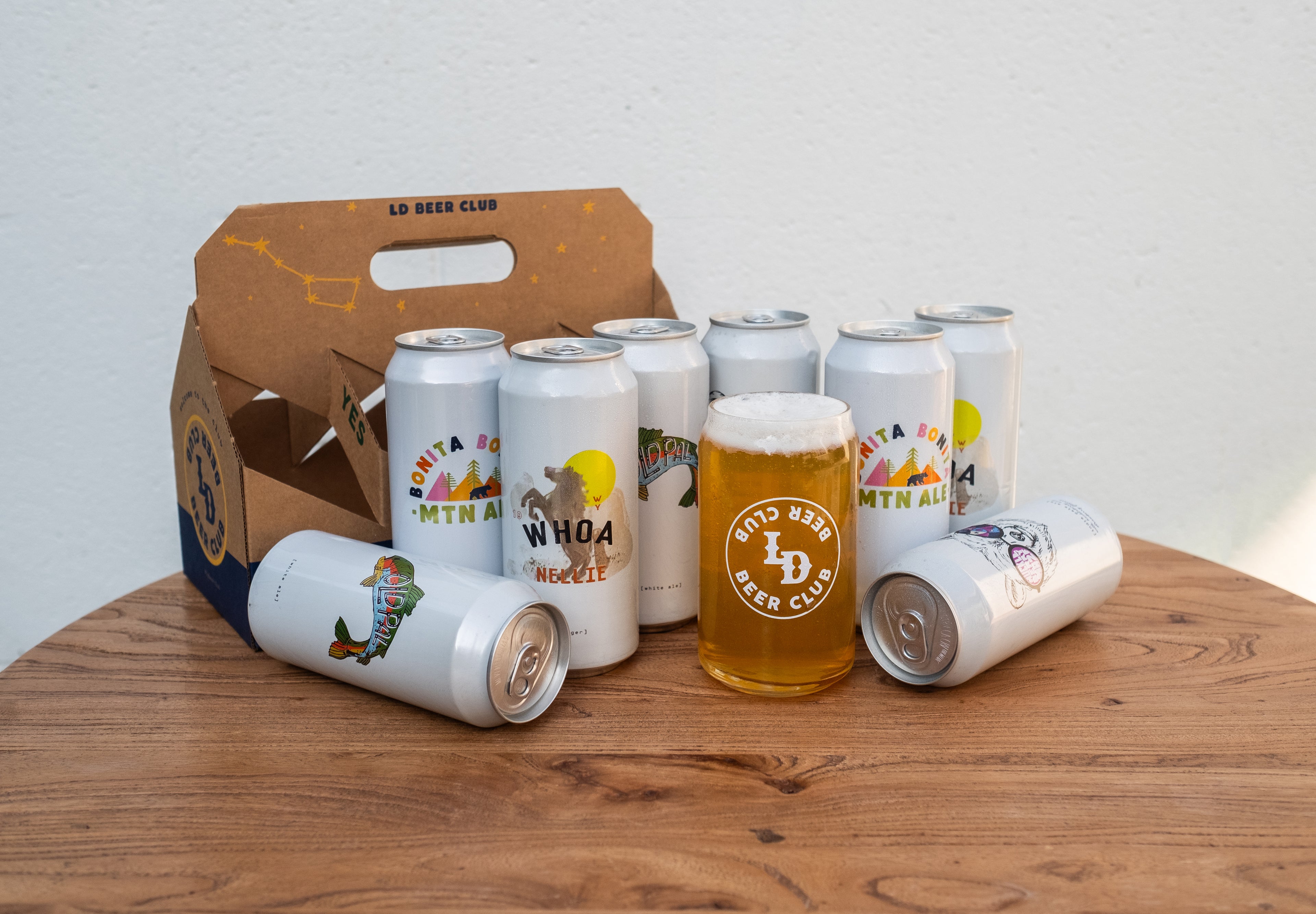 Collection of beer cans and a glass on a wooden table with a branded box.