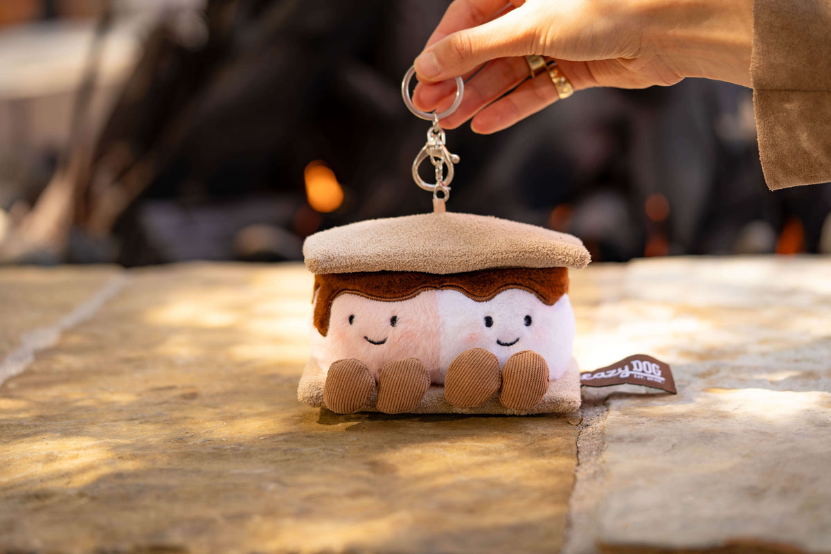 A plush s'mores keychain held by a hand, featuring two smiling marshmallows with brown chocolate-like filling and a beige s'mores hat, set against a warm, blurred outdoor background.